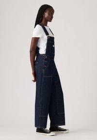 Mujer con trenzas largas vestida con una camiseta blanca, un overol de mezclilla oscuro de pierna ancha y zapatillas negras de caña alta, de pie de perfil.