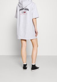 Šedé oversized mikinové šaty s logem na zadní straně, krátkými rukávy a klokankovou kapsou, doplněné černými vysokými teniskami.