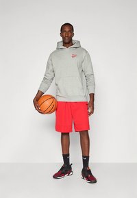 Szürke kapucnis pulóver piros "BULLS" logóval, piros rövidnadrág és fekete sportcipő. Kosárlabdát tart, sima háttér előtt áll.