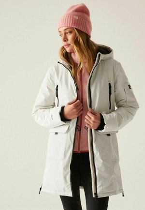 Kobieta ubrana w biały zimowy płaszcz, różową dzianinową czapkę i różową teksturowaną kurtkę, stojąca na jednolitym jasnym tle.