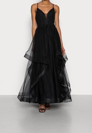 Nainen, joka pukeutunut mustaan hihattomaan iltapukuun, jossa on pitsinen yläosa, kerroksittain laskeutuva läpikuultava hame ja ohuet olkaimet, yhdistettynä mustiin avokauluksellisiin korkokenkiin.