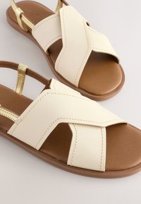 Sandales beige clair avec un design croisé, en cuir synthétique. Semelles intérieures marron et brides dorées pour les accents. Semelle plate, style décontracté.