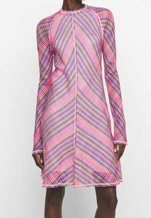 Robe trapèze à manches longues avec un motif à rayures diagonales rose, violettes et orange et coutures à bord brut portée par une personne debout.