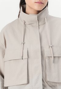 Veste légère beige avec un col montant, deux poches poitrine et des cordons de serrage avec embouts métalliques, présentant une texture douce et lisse.
