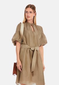 Olivengrünes Leinenkleid mit Puffärmeln, Schlüssel-ausschnitt und gebundenem Taillengürtel. Begleitet von einer kleinen braunen Handtasche.