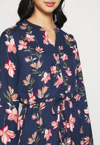 Marineblaues Blumenkleid mit pinken Blumen, Knopfleiste vorne und Bindegürtel. Hergestellt aus weichem Stoff mit langen Ärmeln und kragenloser Ausschnitt.