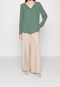 Blouse verte à manches longues avec un col rabattu, associée à un pantalon large beige et décontracté, ainsi qu'à des sandales blanches à brides. Texture de tissu lisse.