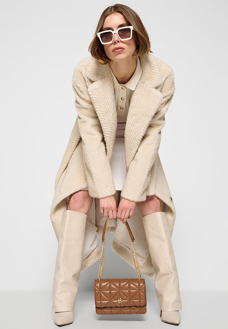 Cappotto beige con texture e ampi risvolti, polo bianca con bottoni, stivali beige al ginocchio e borsa marrone trapuntata con tracolla a catena.