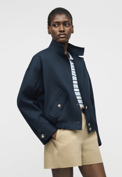 Γυναίκα που φοράει μπουφάν με σκούρο μπλε ψηλό γιακά, ριγέ πουκάμισο και μπεζ σορτς, όρθια με τα χέρια στις τσέπες μπροστά σε απλό φόντο.