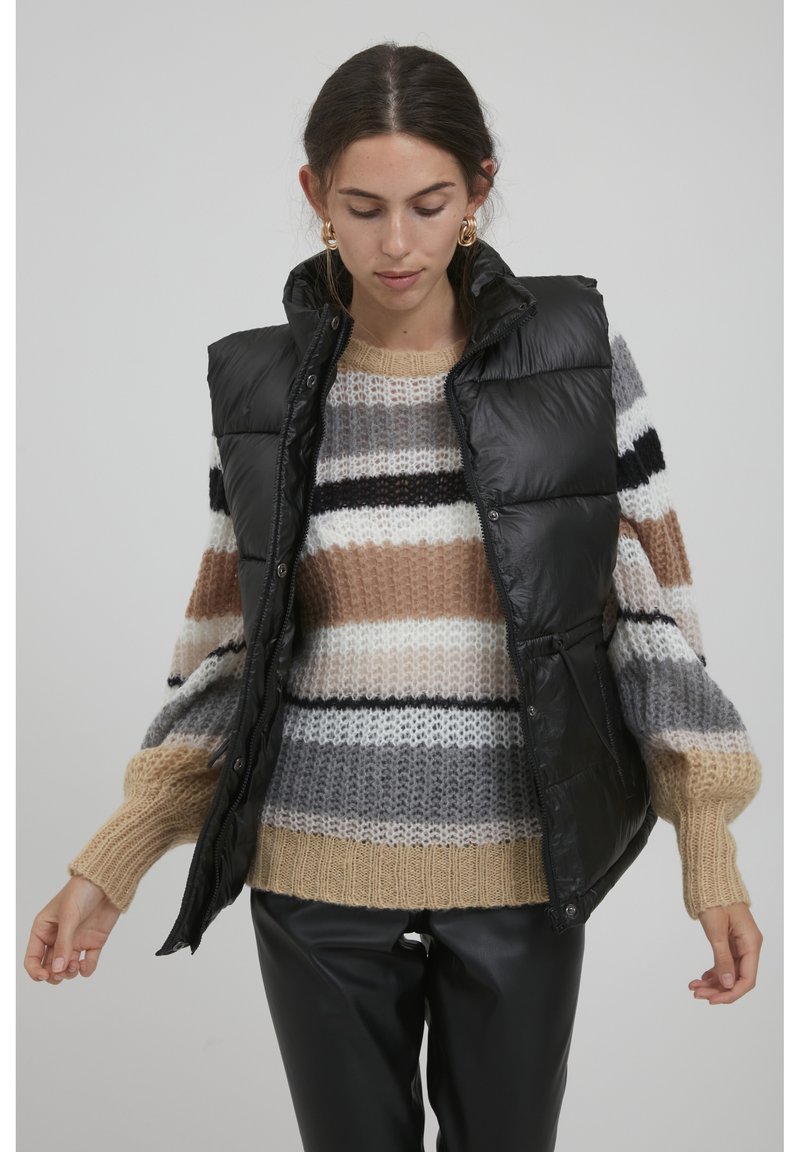 Gilet sans manches en duvet noir avec une texture brillante, poches avant, col haut et fermeture à bouton pression, porté par-dessus un pull en maille rayé.