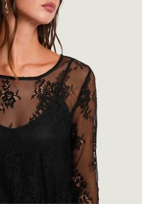 Top de encaje negro de manga larga con un patrón floral transparente, escote redondo y sección inferior sólida en negro. Textura suave y detalles delicados.
