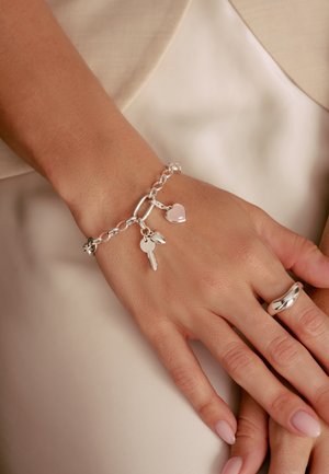 Silberarmband mit großen Gliedern, einem Herzanhänger, einem Schlüsselanhänger und einer polierten Oberfläche. Getragen an einer Hand mit hellem Nagellack.