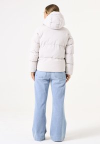 Chaqueta acolchada en gris claro con capucha, diseño acolchado y una parte trasera ligeramente cortada. Combinada con jeans acampanados en azul claro.