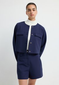 Granatowa krótka kurtka z ozdobnymi złotymi guzikami i dwoma przednimi kieszeniami, w zestawie z dopasowanymi szortami i białym swetrem z golfem.