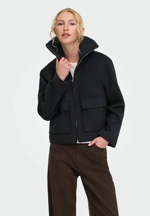 Femme aux cheveux blonds portant une veste noire zippée avec de grandes poches avant et un pantalon marron, posant avec une main près de son col.