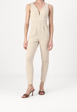 Vrouw draagt een beige mouwloze jumpsuit met een diepe V-hals en bruine hoge hakken met studs, staand op een witte vloer.