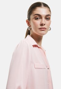 Light pink blouse with a collared neck, front pocket, and button detail. Made of soft fabric with a smooth texture and relaxed fit.