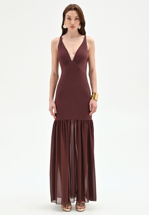 Donna che indossa un abito da sera bordeaux senza maniche con scollo a V profondo e gonna trasparente svasata, braccialetto d'oro, orecchini e sandali con plateau, in piedi frontalmente.