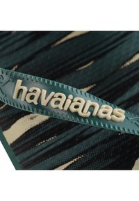 Temno zelena gumijasta sandala s kremno obarvanim logotipom "havaianas", z teksturirano površino in diagonalnim črtastim vzorcem v ozadju.