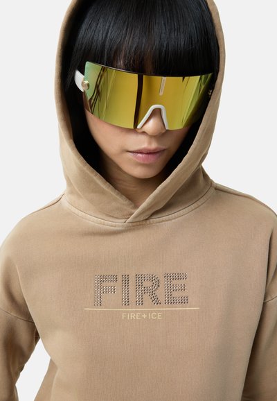 Beżowa bluza z kapturem z napisem "FIRE" ozdobionym cekinami oraz "FIRE+ICE" poniżej. Model nosi duże, sportowe okulary przeciwsłoneczne w złotym odcieniu z białymi akcentami.