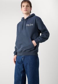 Marineblå hettegenser med frontlomme, snøring i hetten, og "New York" i hvit brodering. Kombinert med løse blå jeans.