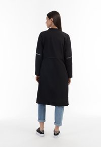 Cappotto lungo nero con chiusura a zip frontale, maniche lunghe e due tasche laterali con zip. Indossato sopra jeans blu con risvolto e sneakers nere.