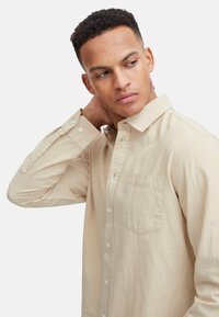 Helles beiges Langarmhemd aus weichem Stoff, mit einem Button-Down-Kragen, einer Brusttasche und weißen Knöpfen auf der Vorderseite.