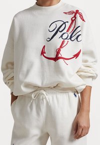 Vit sweatshirt med stort röd och marinblå tryck som visar en ankare och texten "Polo". Avslappnad passform, ribbade ärmslut och mjukt material.