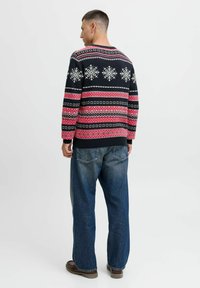 Maglione lavorato a maglia nero e rosso con motivi di fiocchi di neve e disegni geometrici; presenta polsini e orlo a coste, indossato con jeans blu larghi.