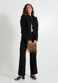 Chaqueta negra corta con cuatro bolsillos frontales y botones plateados, combinada con pantalones negros y un bolso de mano dorado con flecos. Sandalias negras de tacón.