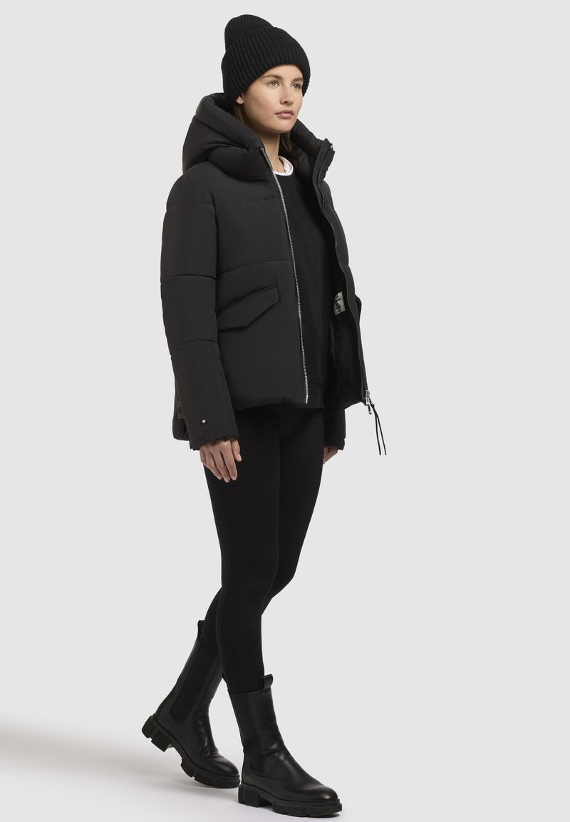 Schwarze Steppjacke mit Kapuze, mit Reißverschluss und Fronttaschen. Trägt man mit schwarzen Leggings und klobigen schwarzen Lederstiefeln.
