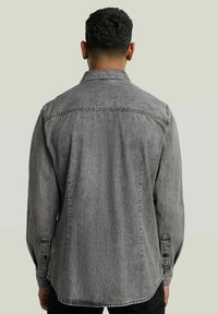Camicia di jeans grigia con maniche lunghe, polsini a bottoni e colletto a punta. Presenta cuciture visibili e una vestibilità rilassata.