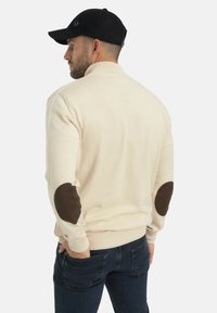 Un homme de profil portant un pull crème avec des patchs noirs aux coudes, un jean noir et une casquette noire, les mains dans les poches arrière.