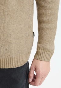 Beige neulepaita, jossa on ribbikuviointi, pitkät hihat ja tiukan istuvuus hihoissa, sekä pieni etiketti sivussa.