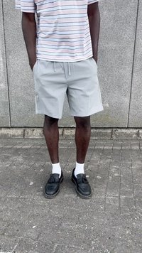 Grey shorts with a drawstring, striped short-sleeve shirt, black loafers, and white socks, standing on a textured pavement.