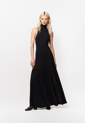 Kobieta o długich blond włosach nosi czarną, bezrękawnikową suknię z golfem i czarne baleriny, stojąc na jednolitym białym tle.