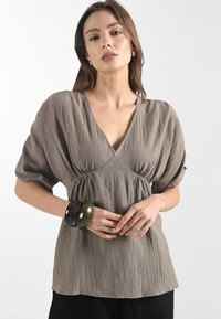 Blusa marrone, con texture, dal profondo scollo a V e maniche a palloncino; indossata con tre grandi bracciali scuri, su uno sfondo chiaro.