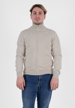 Giacca beige con zip e colletto alto, polsini a coste e orlo. Il tessuto sembra liscio ed elastico. Indossata con jeans in denim blu.