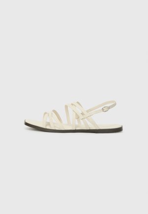 Flad beige sandal med flere tynde stropper og en lille rund sølvspænde på ankelrem mod ensfarvet baggrund.