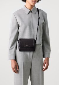 Borsa a tracolla in pelle nera con chiusura a pattina, dettagli in oro e tracolla regolabile, indossata su un outfit a scacchi.
