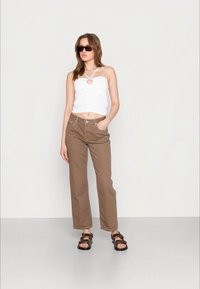 Top cropped branco com detalhe de chave, combinado com calças largas castanhas e sandálias pretas. Modelo com óculos de sol, de pé contra um fundo claro.