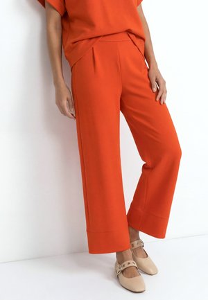 Person wearing wide-leg orange pants and matching top, paired with beige flat shoes with straps and metal eyelets.
