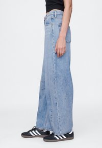 Vaqueros de pierna ancha en denim azul claro con cintura alta, con bolsillos delanteros y traseros, y un bajo sin rematar. Combinados con zapatillas negras.