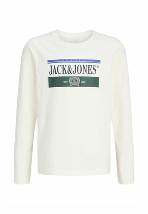 Nastolatek z kręconymi włosami, ubrany w białą, długą koszulkę Jack & Jones, spodnie w kolorze khaki i brązowe buty, stoi i się uśmiecha.