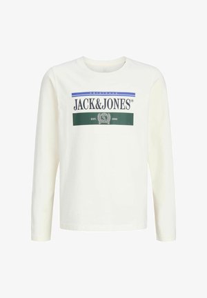 Nastolatek z kręconymi włosami, ubrany w białą, długą koszulkę Jack & Jones, spodnie w kolorze khaki i brązowe buty, stoi i się uśmiecha.