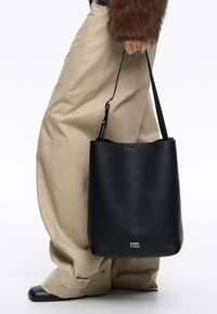 Czarna skórzana torebka typu baniak o teksturowanej powierzchni, solidnym uchwycie i subtelnym logo. Torebka ma minimalistyczny design i zaokrąglony kształt.