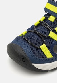 Sapatilhas desportivas azul-marinho com parte superior em malha, detalhes amarelos e atacadores elásticos pretos. Apresentam uma biqueira em borracha preta texturizada e um símbolo de aviso.