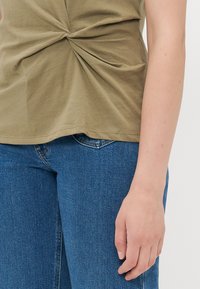Olivenfarget, kortermet topp med vridd frontdetalj, kombinert med blå denimjeans. Glatt bomullsstoff, avslappet passform.