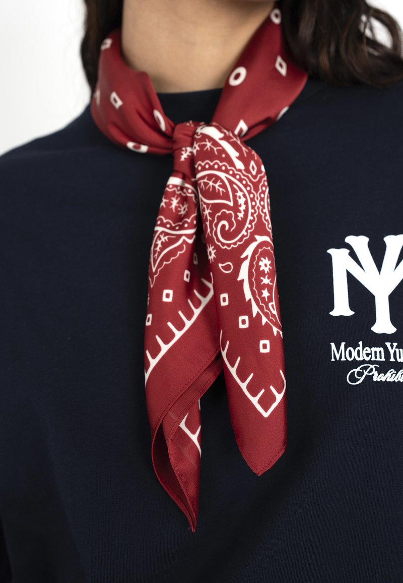 Mujer que lleva un pañuelo rojo con estampado de cachemira atado alrededor de su cuello y una camisa azul marino con texto blanco en el pecho.