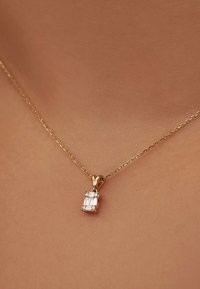 Goldene Halskette mit feiner Kette, versehen mit einem rechteckigen Anhänger, der mit klaren, rechteckig geschliffenen Steinen verziert ist und auf der Haut ruht.